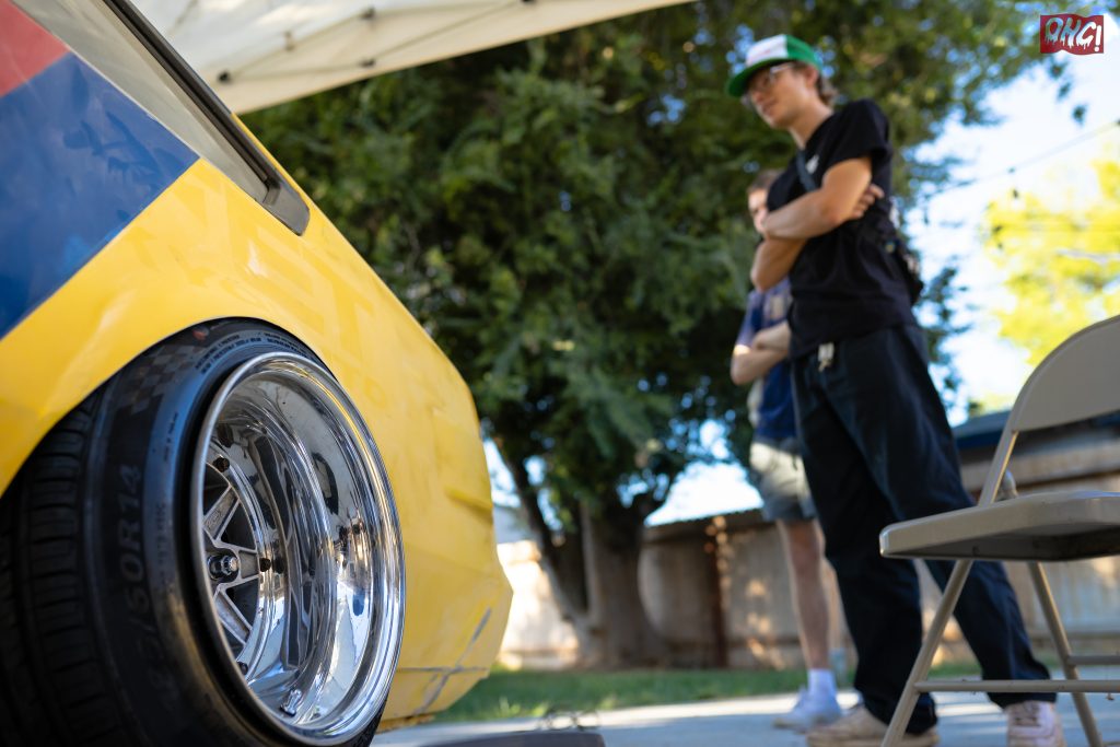 Close-up of a yellow car’s polished rim and tire with two people in the background at an outdoor event.