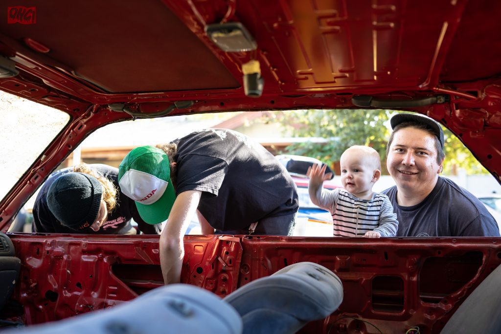 Family bonding while working on a red car interior. Dad holding baby, two men in green and black hats fixing the car.