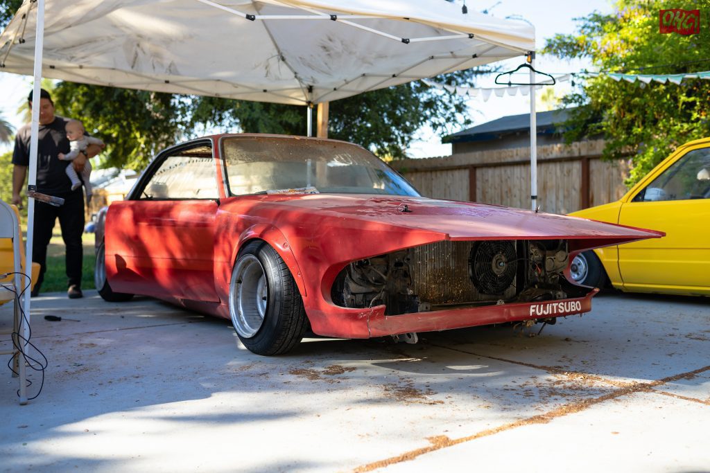 Red vintage car under white tent, partial restoration, with Fujitsubo part, baby and adult in the background.