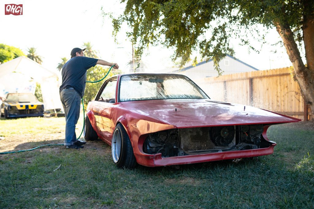 Person washing a red vintage car's stripped frame with a hose in a sunny backyard, OHG! logo in the corner.