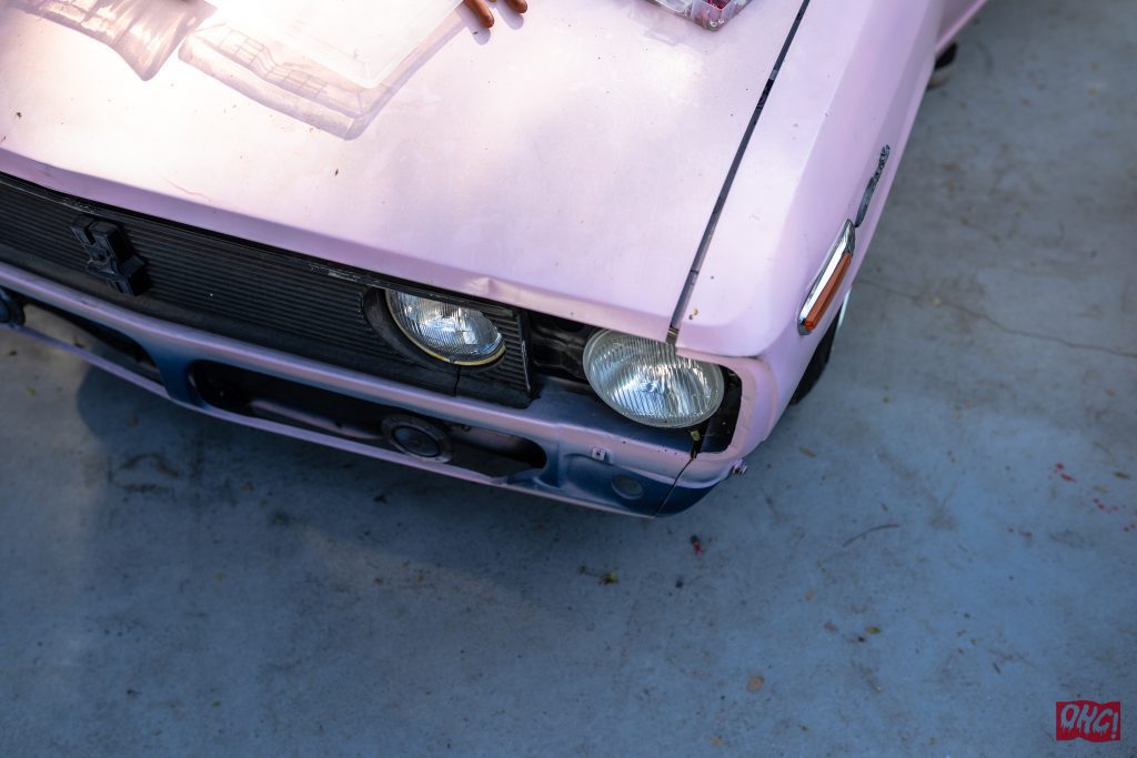 Close-up of a vintage pink car's front with noticeable headlight and grille. OHGI Japanese lettering visible on ground.