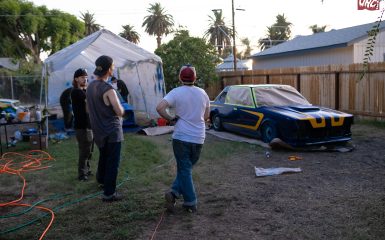 People painting custom race car outdoors in a backyard garage setup with tarp shelter and painting equipment.