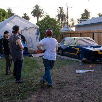 People painting custom race car outdoors in a backyard garage setup with tarp shelter and painting equipment.