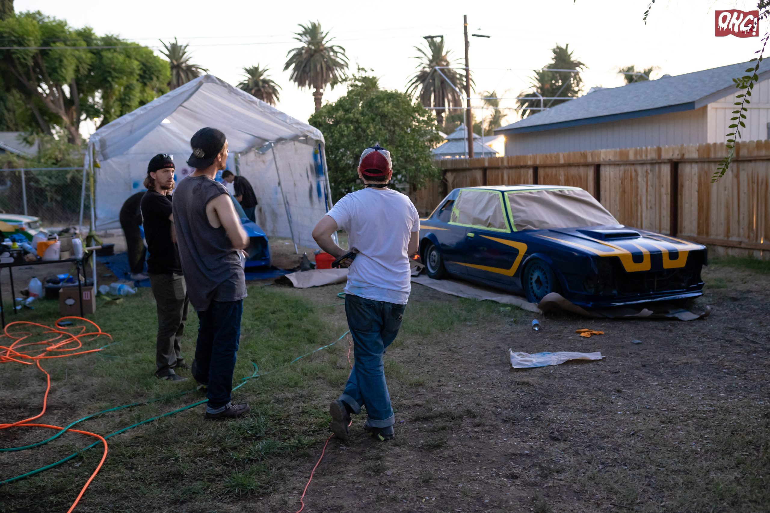 People painting custom race car outdoors in a backyard garage setup with tarp shelter and painting equipment.