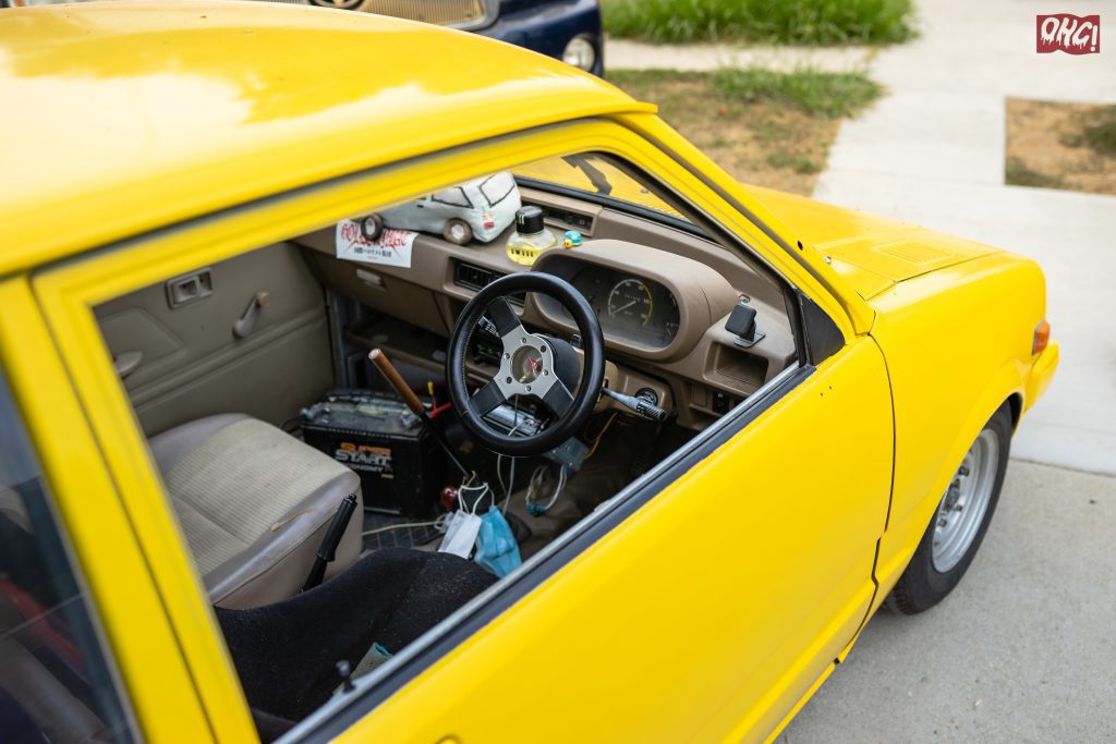 Interior view of a classic yellow car showing the dashboard, steering wheel, and seats, with a START battery on the floor.