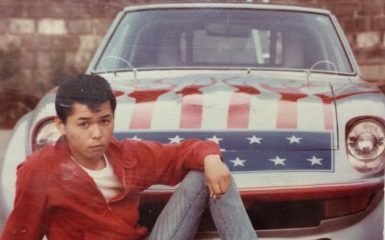 Young man in red jacket poses in front of an American flag-themed car with red flames, white stars, and a blue background.
