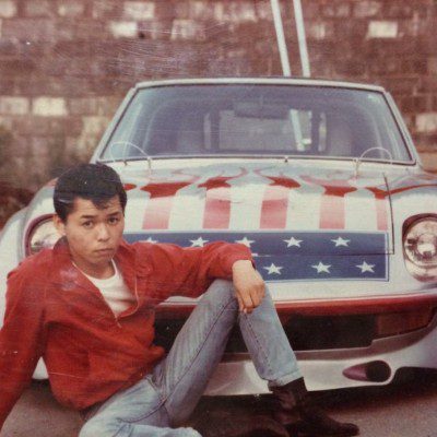 Young man in red jacket poses in front of an American flag-themed car with red flames, white stars, and a blue background.