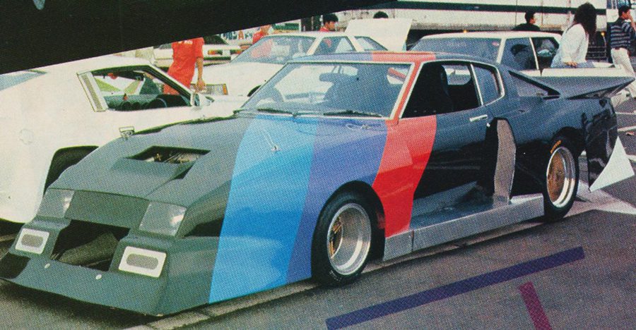 Vibrant custom race car at an outdoor event, featuring a tricolor paint scheme of blue, red, and black. Classic cars in background.