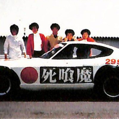 A white sports car with Shinigami and 2998cc written on it, and six people posing behind it on a sunny day.