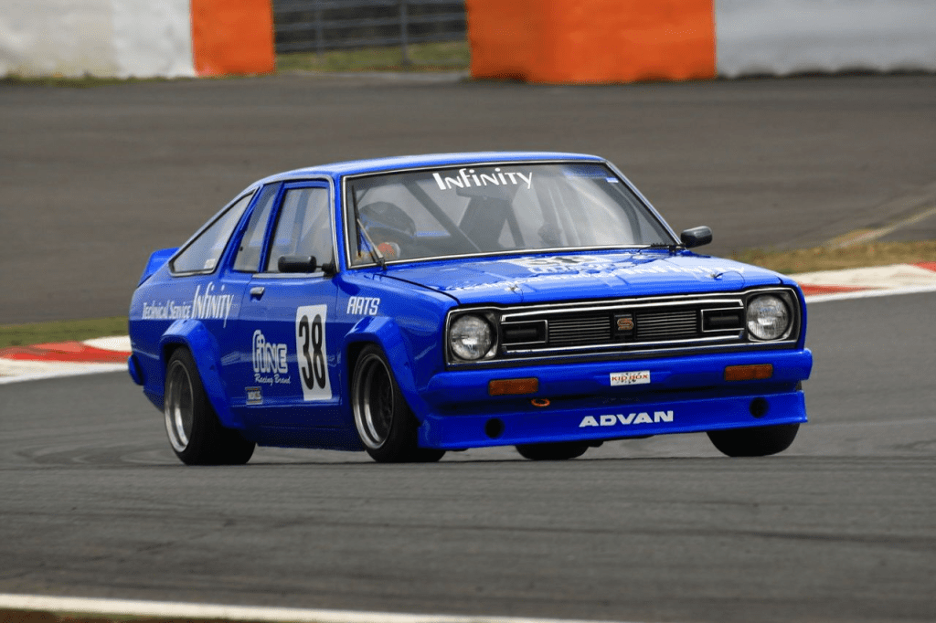 Blue racing car number 38 with Infinity branding, fine racing team, drifting at a track; view from the front-left.