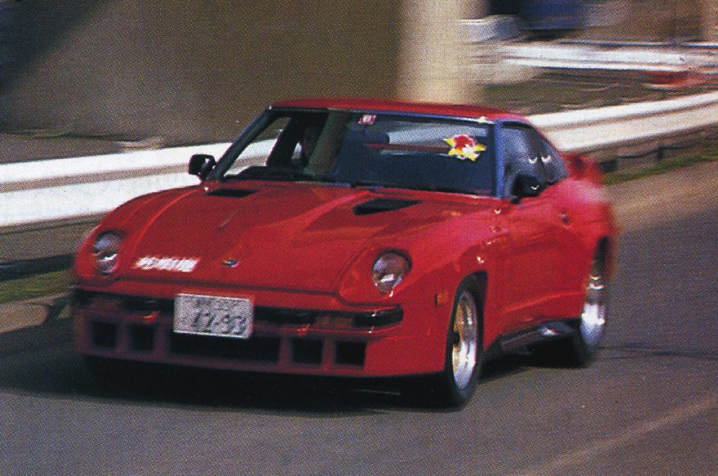 Red classic sporty car with Japanese license plate number '7953,' driving on a road near guardrails.
