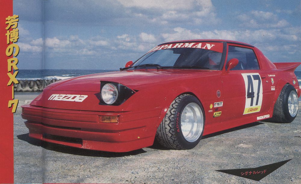 Red Mazda RX-7 race car with a 47 decal, parked near the ocean under a blue sky. Vertical Japanese text on the left.