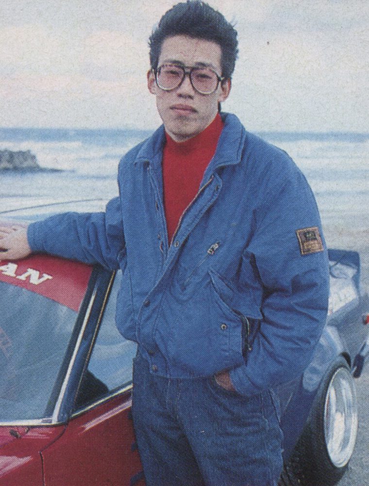 A man in glasses and a blue jacket leans on a red Nissan sports car by the beach, ocean waves in the background.