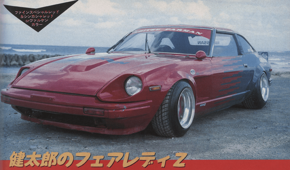 Modified Nissan Fairlady Z in red with racing decals, parked seaside. Japanese text reads Kentaro's Fairlady Z.