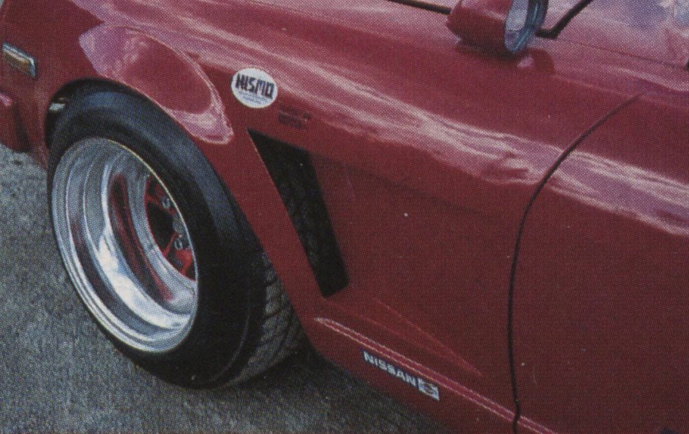 Close-up of a red Nissan car's fender showcasing NISMO badge and deep dish wheels.