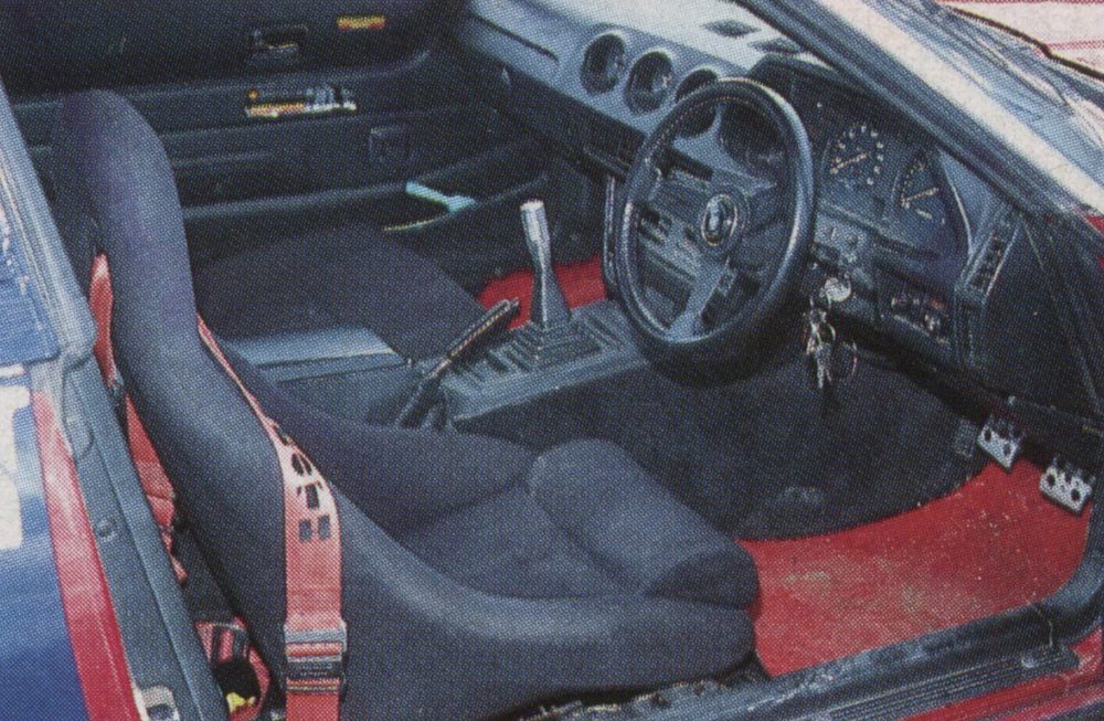 Interior of a sports car with red racing harness and red floor mats, featuring a manual transmission and sporty dashboard.