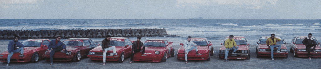 A row of red sports cars with men posing in front, by the ocean, each car's windshield displaying the word Garnet.