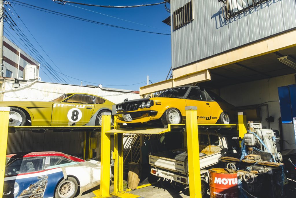 Vintage yellow and green cars parked on a mechanical lift in an industrial Japanese garage under clear blue sky.