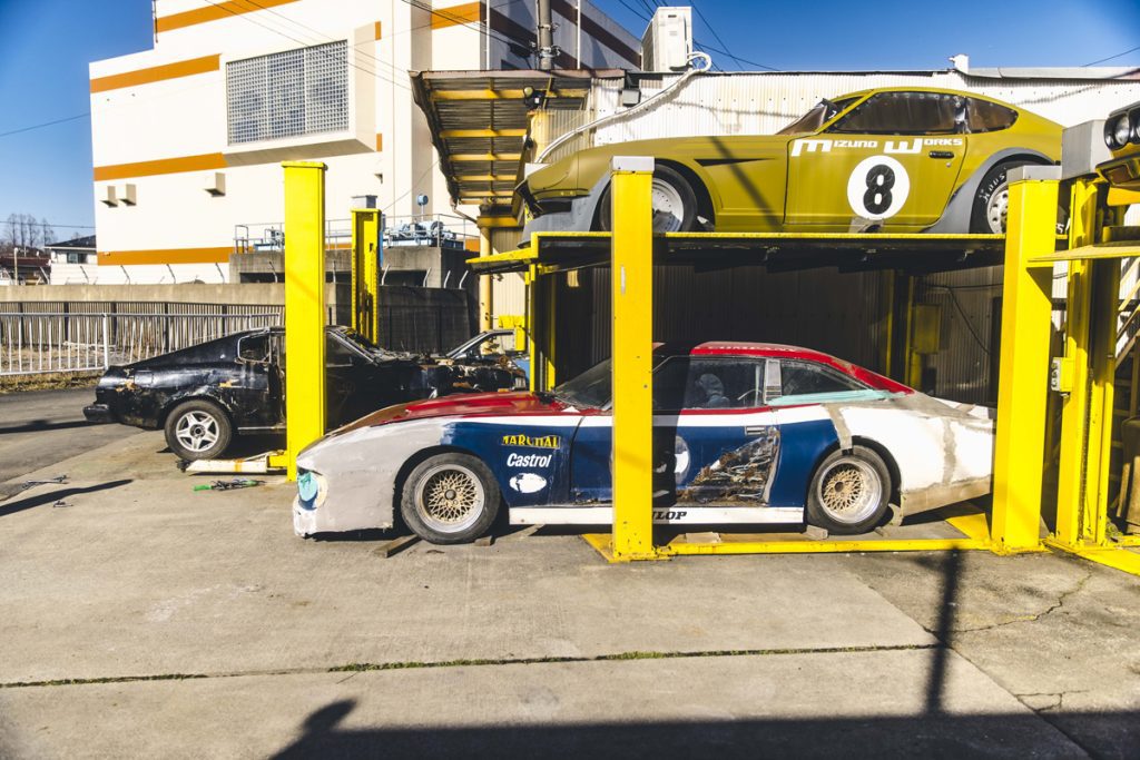 Three classic sports cars parked in a garage with yellow lifts, including a green No. 8 Mizuno Works car on the top level.