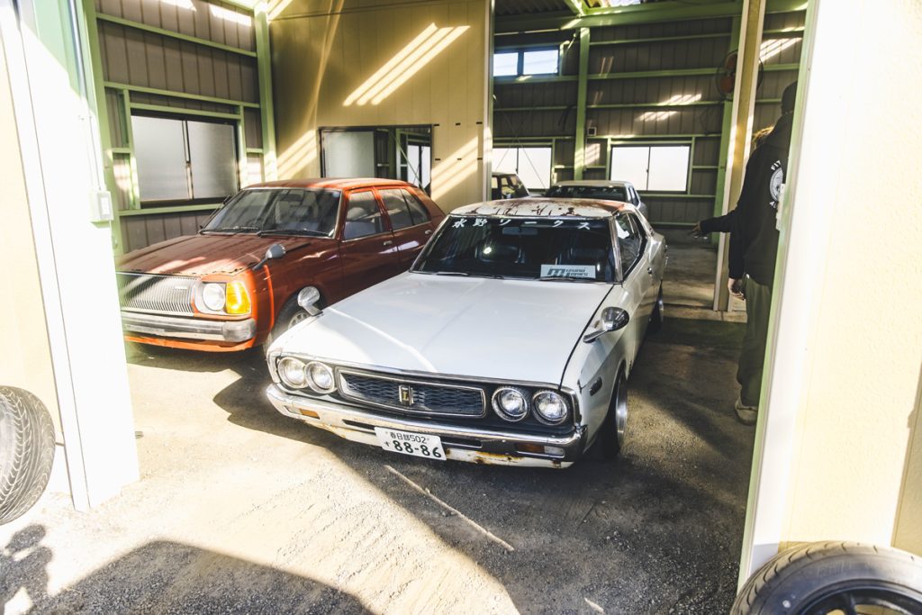 Two classic Japanese cars, a red and a white one, parked in a garage with sunlight streaming in through the windows.