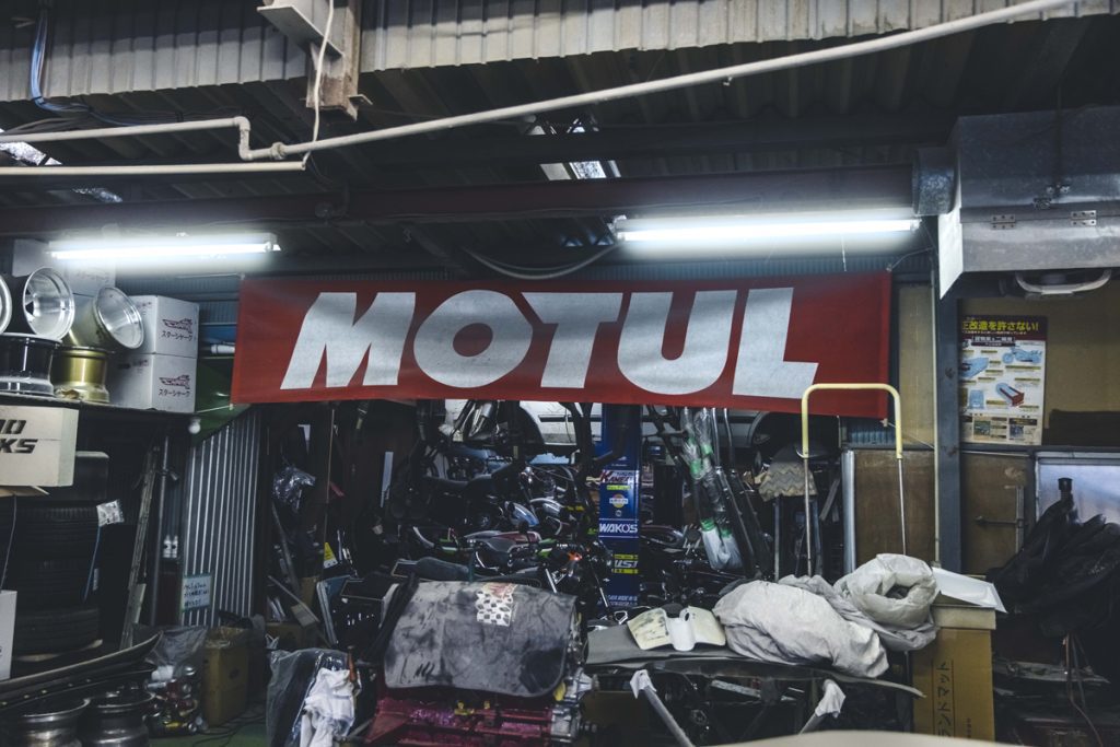 Motul banner inside a cluttered garage with car parts and tires, Japanese signs, and vehicle equipment.