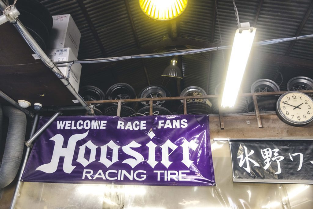 Hoosier Racing Tire sign in a repair shop with rim tires, ceiling lights, clock, and Japanese sign '氷野ワ'.Translation:氷 (kōri) - ice
野 (no) - fieldThe above text may be correct but not certainUsually a phrase has a proper meaning. This