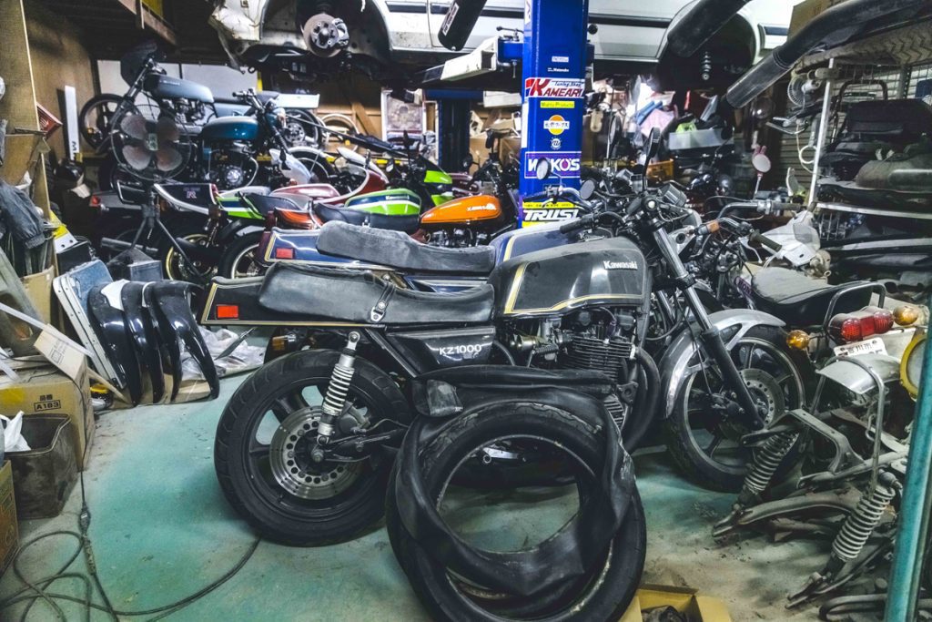 Vintage motorcycles and spare parts in a cluttered workshop, featuring a Kawasaki KZ1000 and various other bikes.