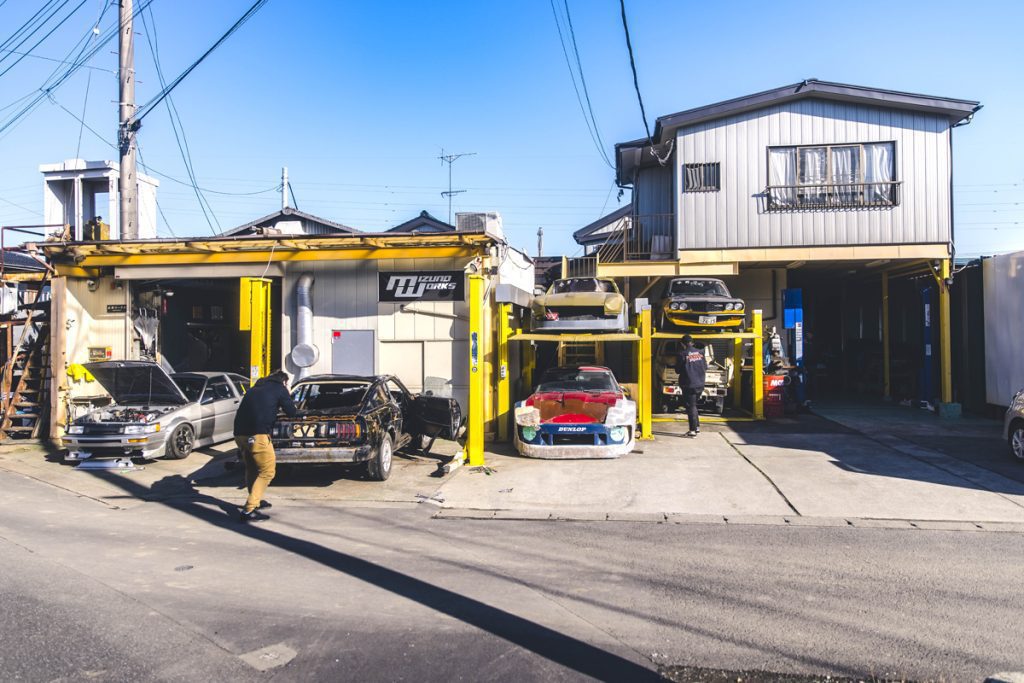 Japanese auto repair shop, featuring classic cars with custom modifications, labeled Izumo Works, showcasing high-level automotive maintenance.