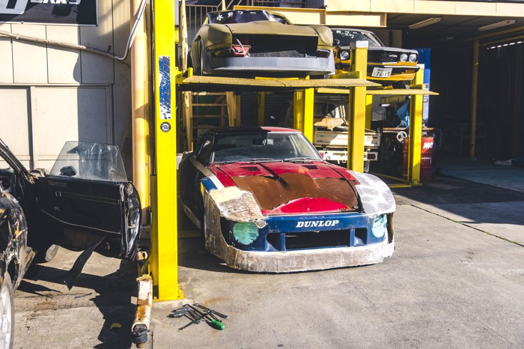 Old racing cars undergoing restoration at an auto workshop with a yellow car lift and tools in the foreground.