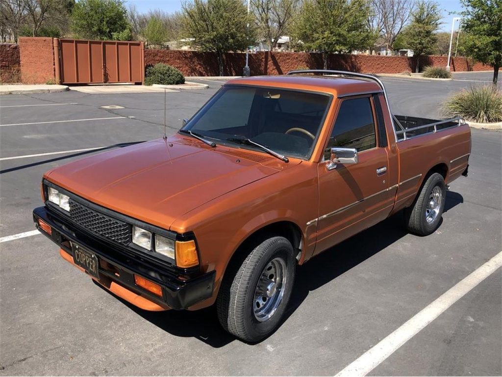 Vintage copper-colored pickup truck parked in a lot with trees and a brick wall in the background.