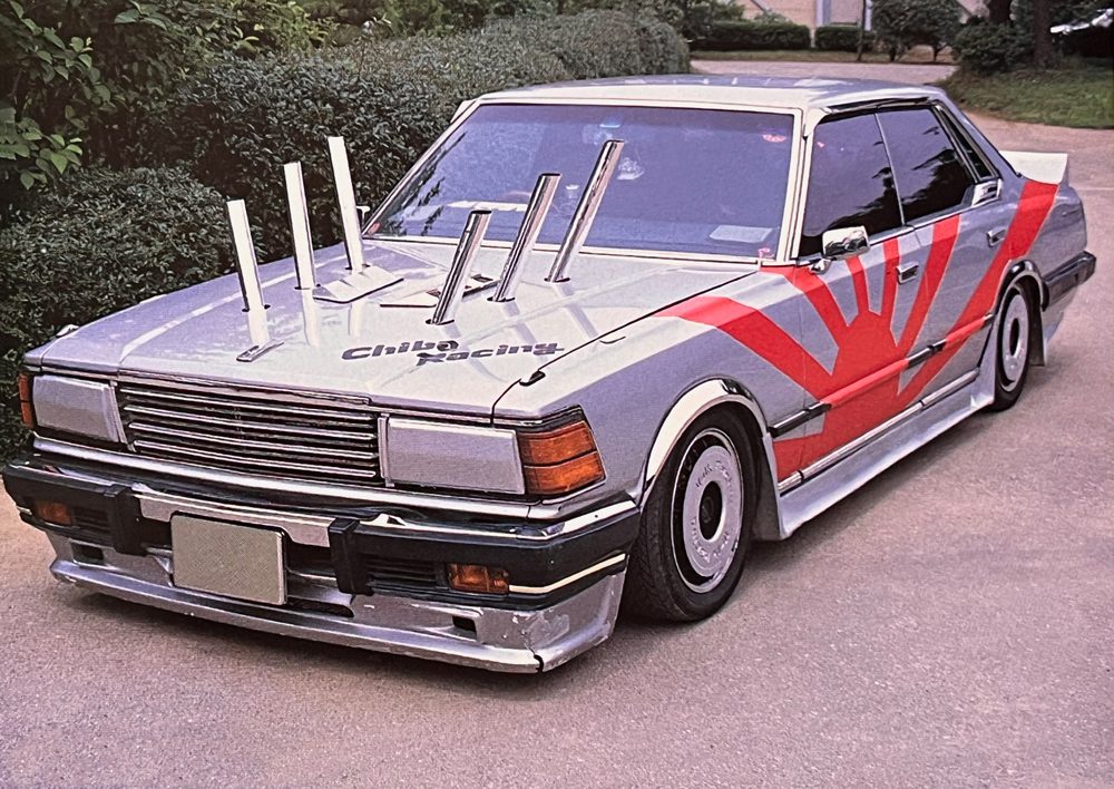 Modified car with Chiba Racing logo, custom exhaust pipes, and rising sun design on the side, parked on a driveway.