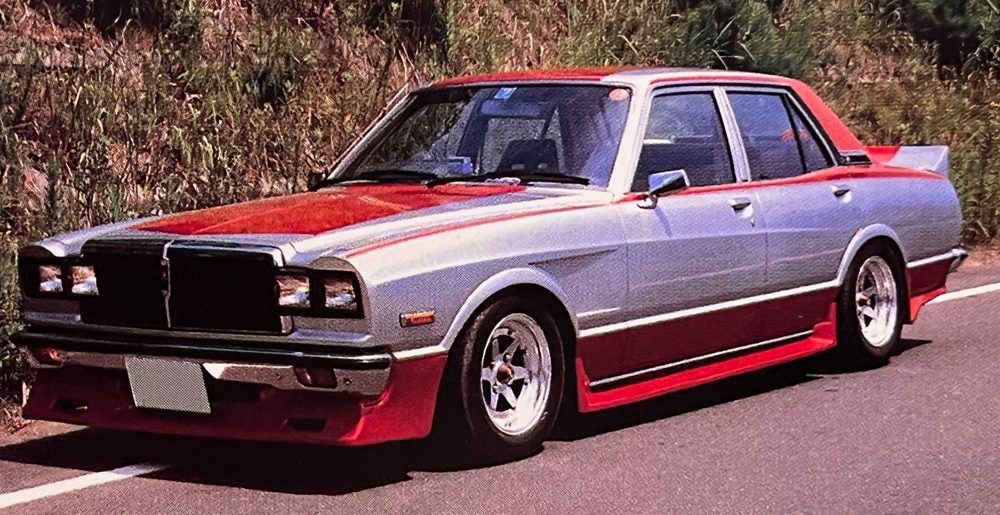 Vintage modified Japanese car with red and silver paint, aftermarket wheels, and a custom front bumper on a scenic road.