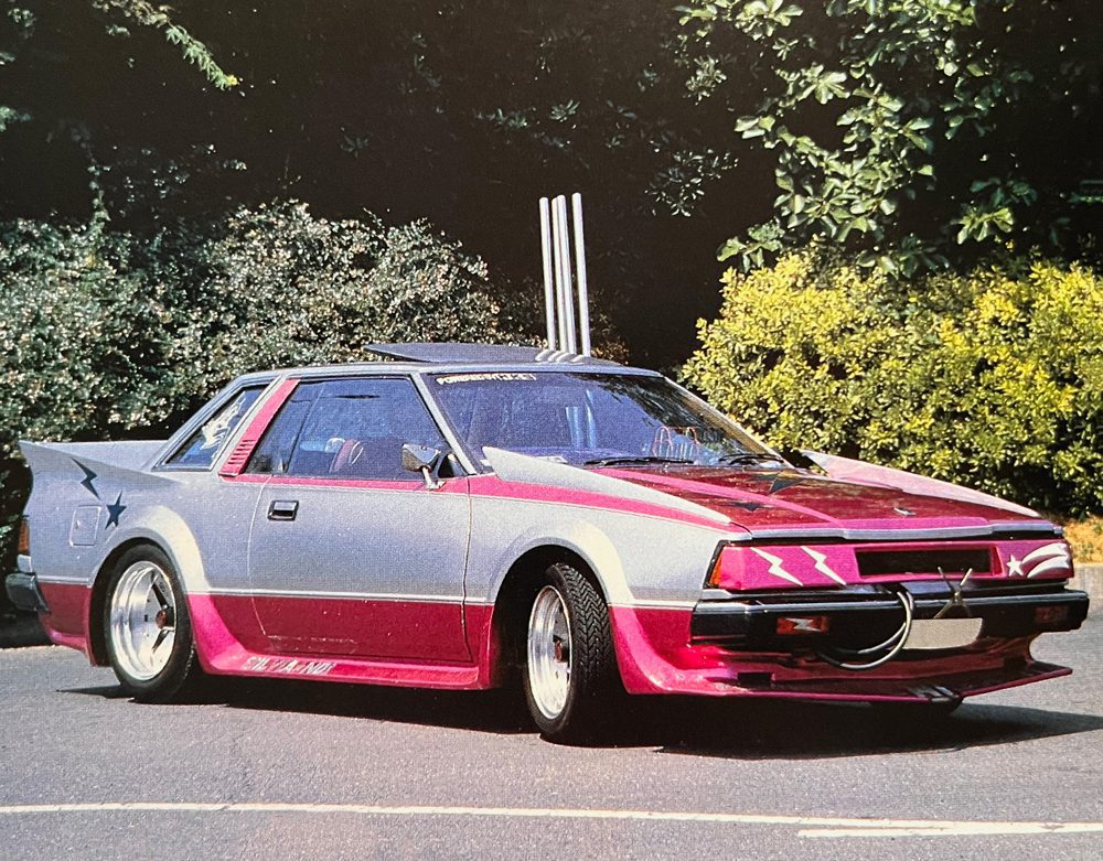 Custom retro car with unique colors and design, featuring Japanese text on the side against a lush green background.