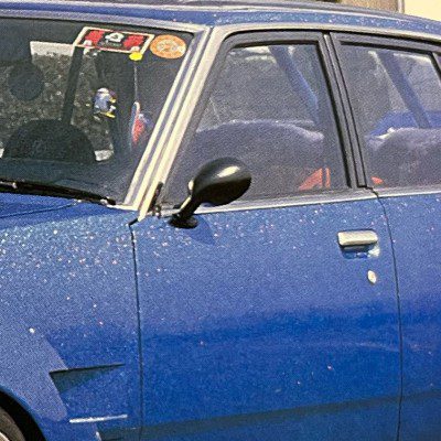 Vintage blue car with Japanese stickers on windshield, showcasing classic design and detailed exterior, captured in daylight.