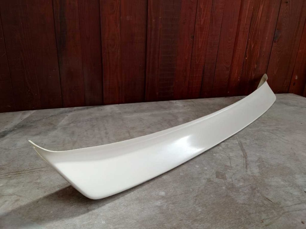 White car spoiler displayed on concrete floor with wooden wall background. Perfect for enhancing vehicle aerodynamics.