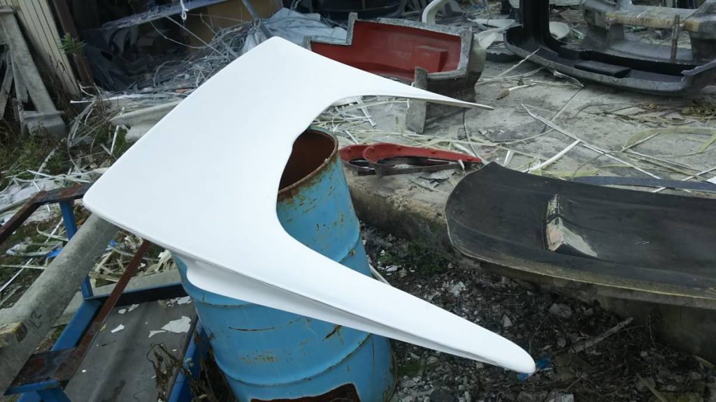 A white car spoiler sits on a rusty blue barrel in a cluttered workshop, ready for installation.