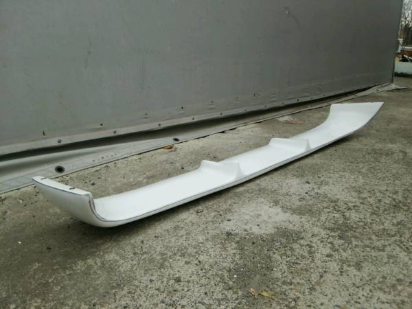 White car rear bumper spoiler on a concrete surface with a metal wall background.