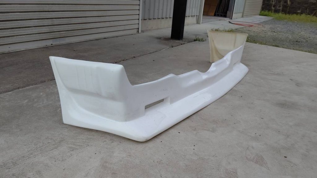 White front bumper for a car placed on concrete ground near a metallic building garage.