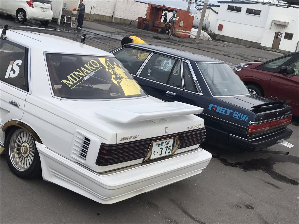 Two custom-tuned Toyota Mark II cars parked in a lot, featuring MINAKO and RET decals and distinct aerodynamic kits.