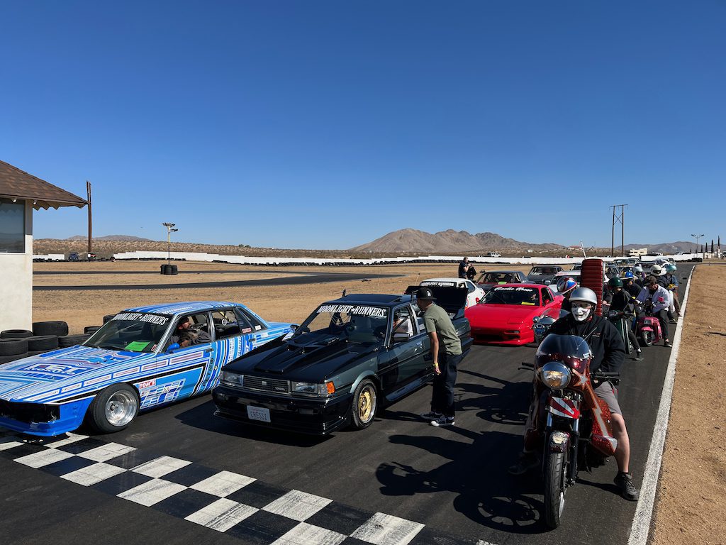 Line of drift cars and motorcycles at a racetrack with people gathered under clear blue sky, mountains in the background.