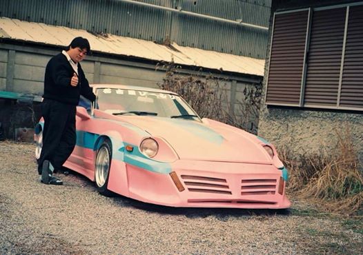 Man gives thumbs up next to customized pink sports car with blue accents, parked outside warehouse; Japanese text on windshield.