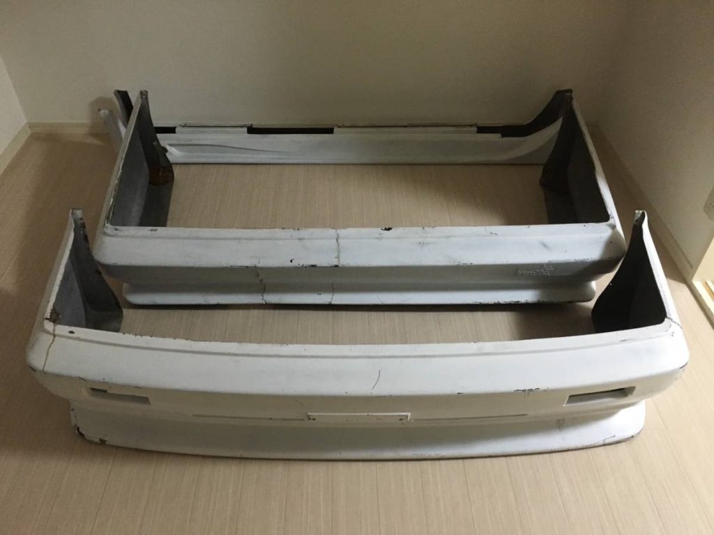 Two used white car bumpers are placed on a light wooden floor, showing signs of wear and minor cracks.