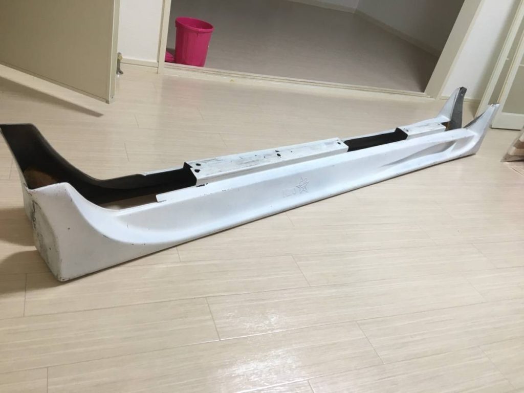 White car bumper with black accent sitting on beige wooden floor next to an open door and pink bucket in the background.
