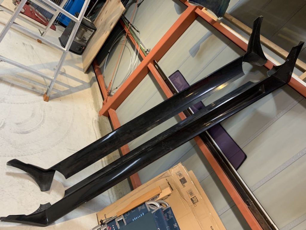 Black car side skirts in an automotive garage, with a ladder and various tools in the background.