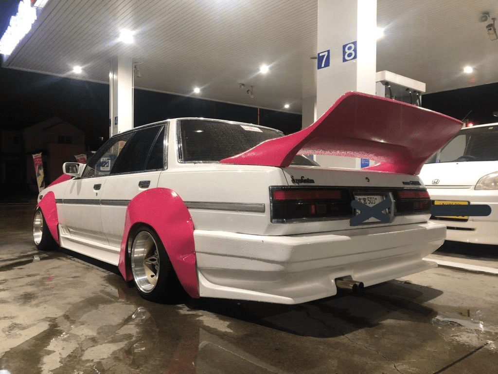 Modified white sedan with pink fender flares and spoiler at a gas station at night in Japan, license plate obscured.