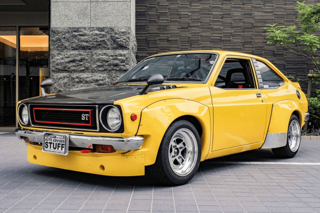 Vintage yellow classic car with custom modifications and ST badge, parked outside a building. License plate reads STUFF.