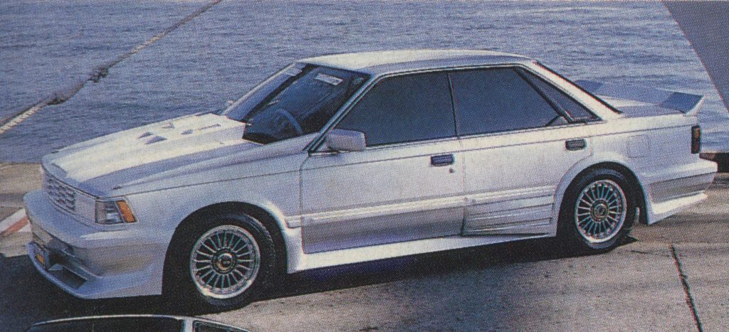 Modified vintage white Japanese sedan with custom body kit and sport rims parked by the water.