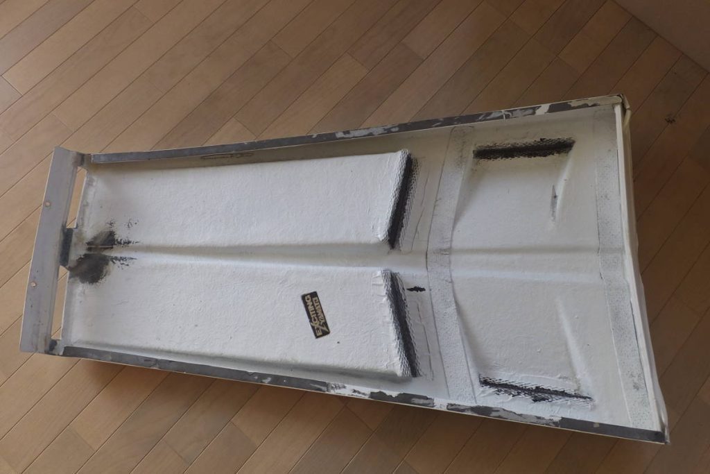Used metal part with cladding sticker in Japanese on a wooden floor, showing signs of wear and damage.