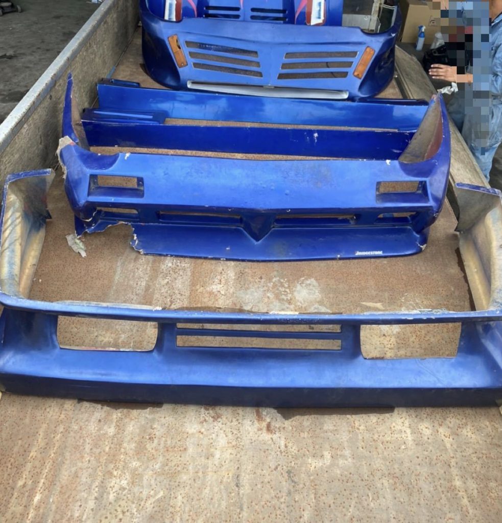 Blue car bumpers in a truck bed, ready for transport or installation. Some parts have minor damage. Bridgestone logo visible.