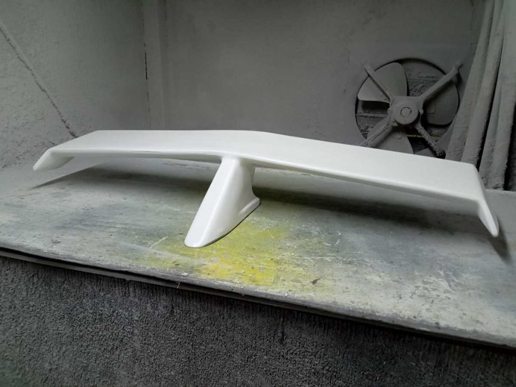 White car spoiler in a workshop with a ventilator in the background, on a dusty workbench.